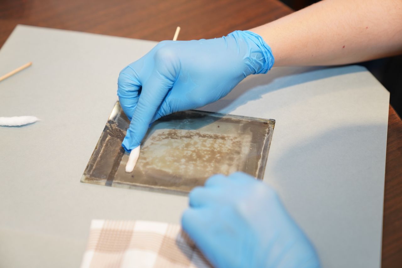 Hands in gloves carefully restoring vintage artwork on glass using cleaning tools.
